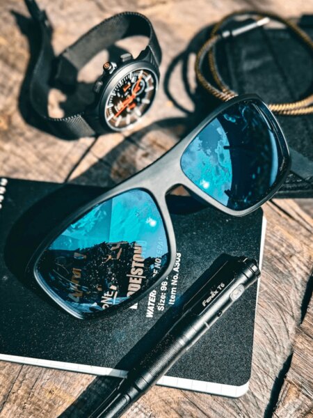 Polarisierte Sonnenbrille von Wiley X auf dem Tisch mit einer Uhr, einem taktischen Notizbuch und einer Taschenlampe