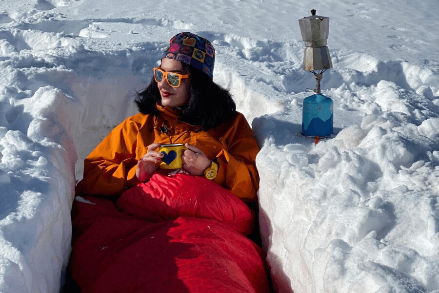 Eine Frau mit einem Blechbecher sitzt in einer Grube in einem Winterschlafsack.