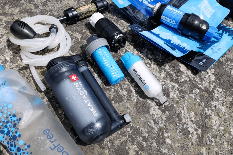 Ein Satz verschiedener tragbarer Wasserfilter von Sawyer, HydraPak und Katadyn auf steinernem Untergrund. Foto: Rigad