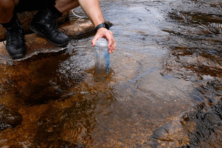 Eine Person schöpft Wasser aus einem Bach in eine weiche Flasche mit Squeeze-Filter bei einem Outdoor-Ausflug. Foto: Rigad
