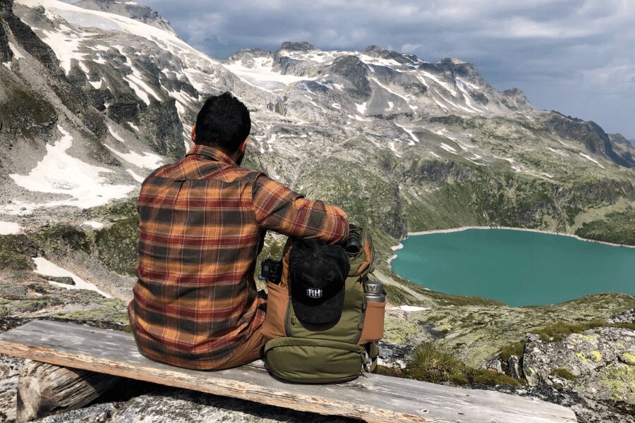 Mann mit Rucksack sitzt in den Bergen und schaut auf den See. Quelle: Rigad