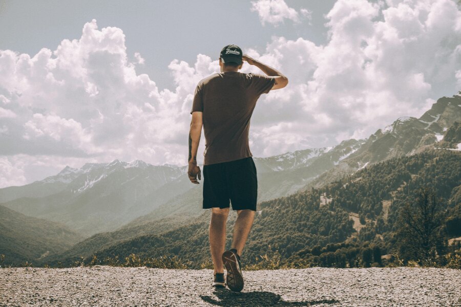 Ein Mann in einem T-Shirt und Shorts steht mit dem Rücken zur Kamera mit Blick auf die Berglandschaft.