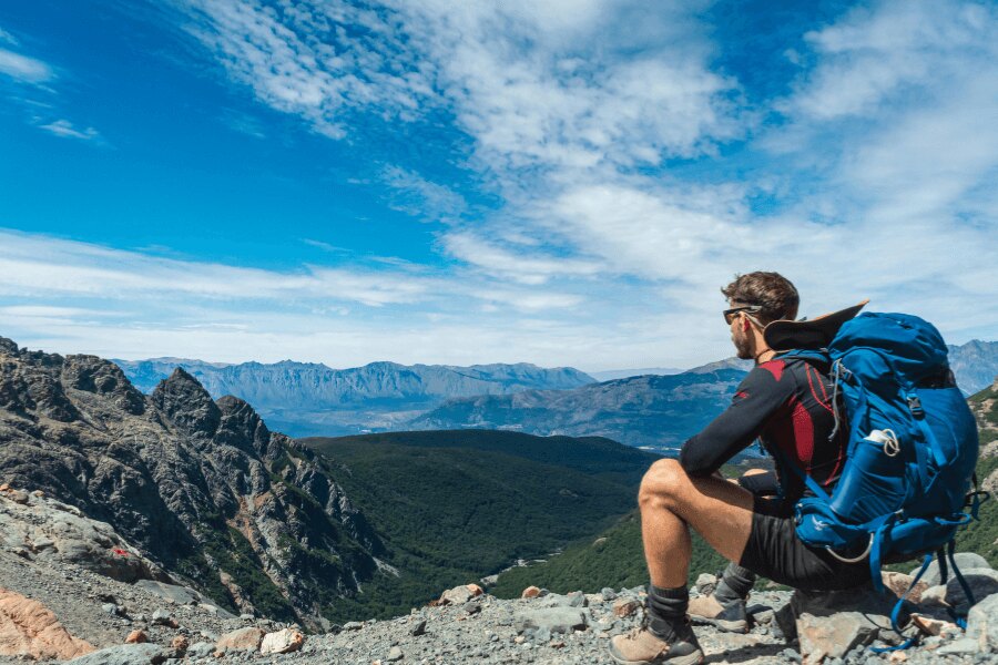 Ein Mann sitzt auf einem Felsen mit einem Rucksack und funktionaler Kleidung und beobachtet die Berglandschaft. Foto: https://www.pexels.com/cs-cz/foto/krajina-hory-priroda-muz-15954827/