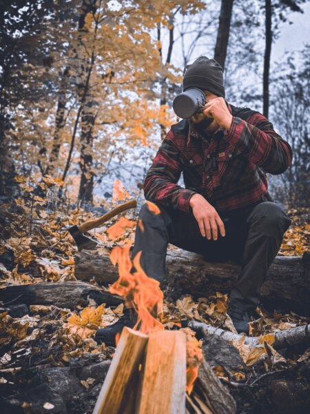 Ein Mann in einer karierten Jacke sitzt am Feuer im herbstlichen Wald und trinkt aus einem Metallbecher. Herbstliches Camping mit dem Duft von Holz und der Ruhe der Natur. Foto: Rigad