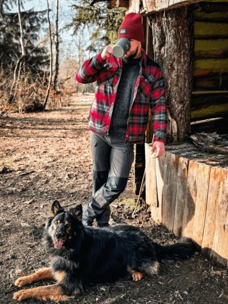 Ein Mann in roter Jacke und Mütze trinkt aus einer Tasse an einer Holzhütte, ein Hund liegt zu seinen Füßen. Herbstcamping mit natürlichem Komfort und treuem Begleiter. Foto: Rigad