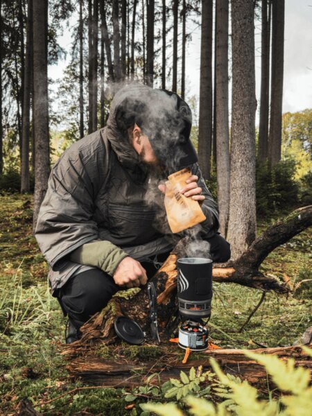 Ein Mann in einer gefütterten Jacke kniet im Wald bei einem Jetboil-Kocher, aus dem Dampf aufsteigt. Er bereitet warmes Essen oder ein Getränk als Energie- und Wärmequelle während eines Herbstausflugs zu. Foto: Rigad