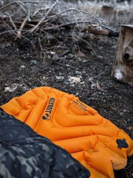 Klymit Insulated Static V Lite Isomatte in orangener Farbe am Boden neben der Feuerstelle, teilweise von einem Schlafsack mit Tarnmuster bedeckt. Isolation gegen Kälte und Feuchtigkeit beim Schlafen im Freien. Foto: Rigad