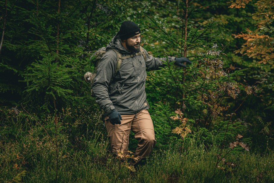 Ein Mann im herbstlichen Wald in einer grauen Winterjacke Yukon mit Climashield™ Apex<sup>®</sup> Isolierung von Helikon-Tex<sup>®</sup>, Mütze und Handschuhen, mit Rucksack; eine Demonstration des richtigen Schichtens und der schützenden Außenschicht gegen Nässe und Wind. Foto: Rigad