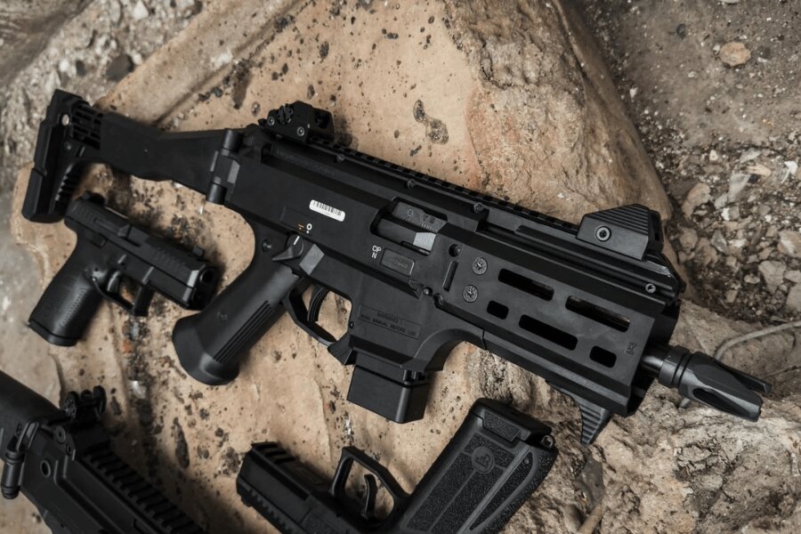 Das moderne Maschinengewehr CZ Scorpion EVO 3 mit einem Reflexvisier und einer Teleskopschulterstütze liegt auf dem Boden neben Pistolen und Magazinen. Foto: Rigad