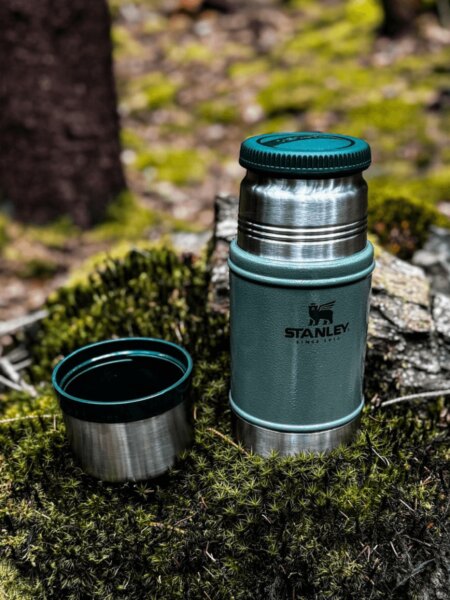 Offene Stanley-Essens-Thermoskanne mit einem Becher auf Moos im Wald platziert.